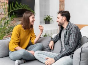 A couple sit on a gray couch, engaged in conversation in a modern, bright living room.