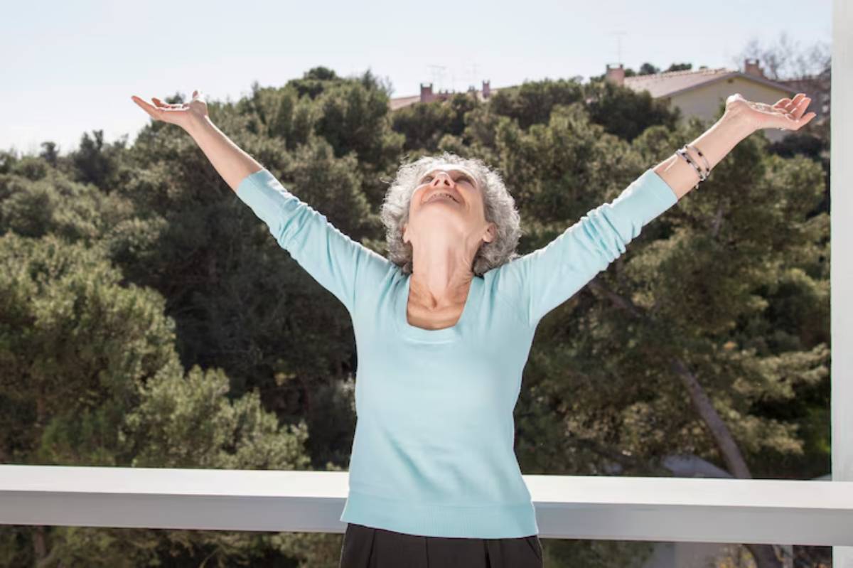 An elderly woman stands on a balcony with arms raised triumphantly against a backdrop of greenery and a clear blue sky.