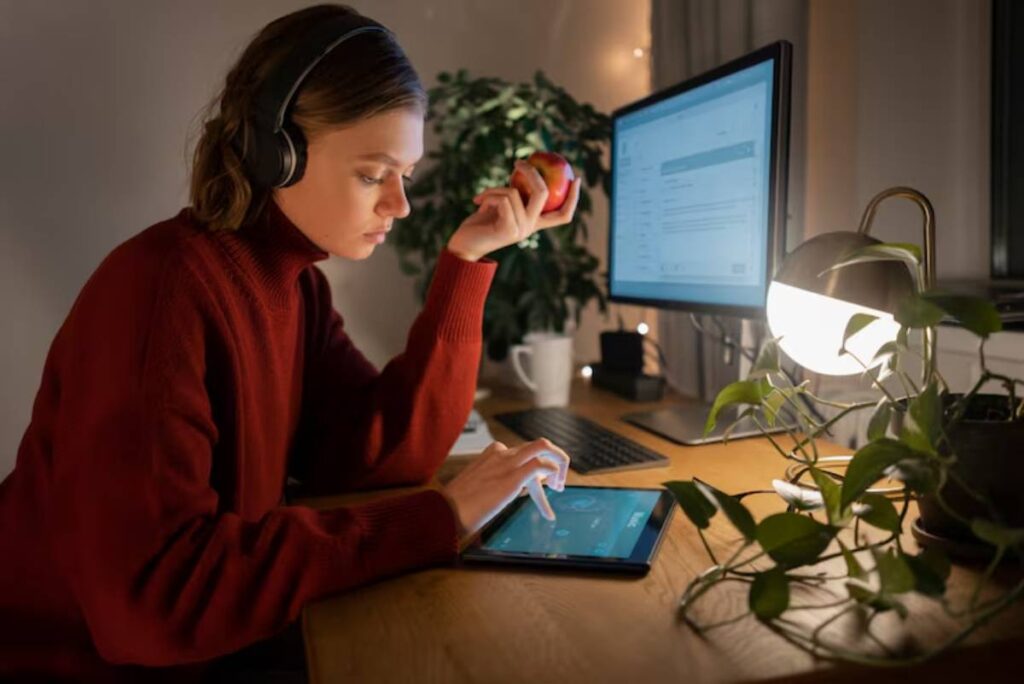 A person in a cozy sweater sits at a desk, using a tablet while holding an apple. A warm lamp and plants enhance the workspace ambiance.
