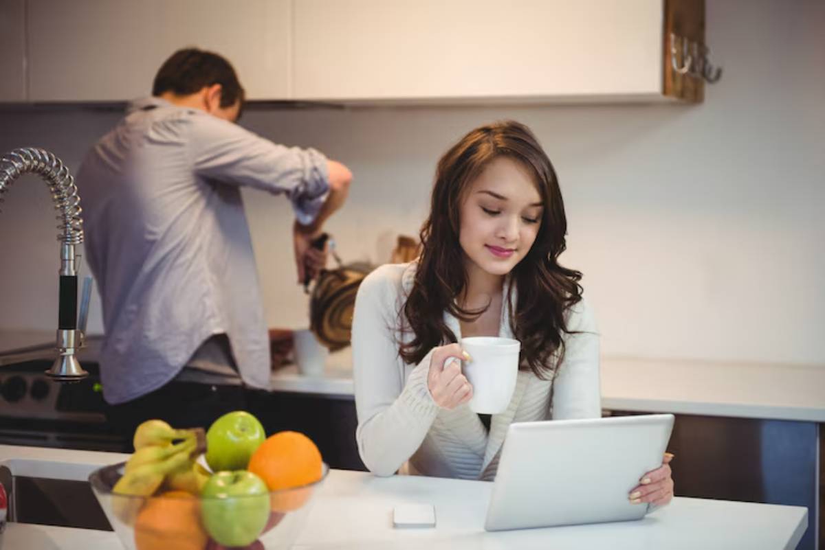 A woman sits at a kitchen counter, holding a cup, using a tablet, while a person prepares food in the background. Fresh fruit is visible.