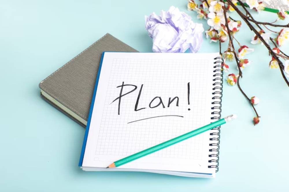 A notebook with 'Plan!' written in bold on grid paper, surrounded by a pencil, crumpled paper, and decorative flowers.