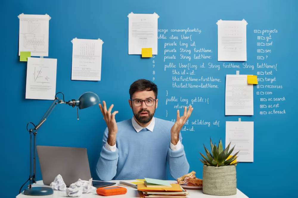 A person in a blue sweater gestures expressively while sitting at a desk cluttered with papers, a laptop, and a potted plant against a blue wall.