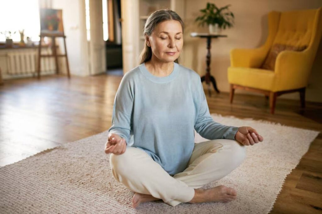 A person meditating on a soft rug in a cozy, well-lit living room, surrounded by plants and a yellow armchair.