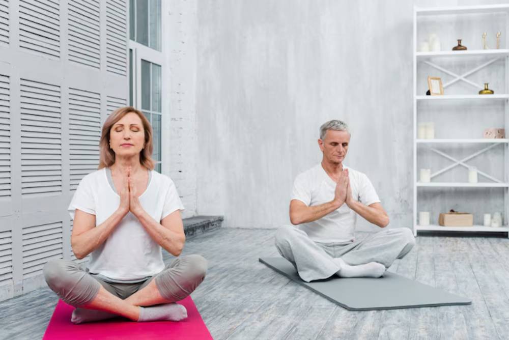 A woman and a man meditate in a serene, minimalist space, sitting cross-legged on yoga mats, hands in prayer position.