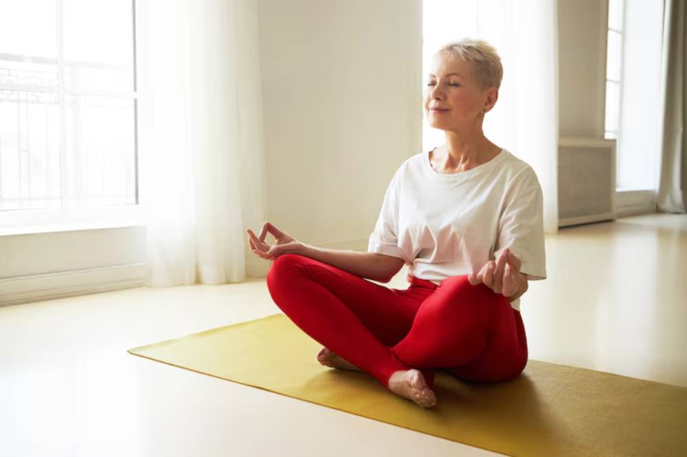 A person practices yoga in a serene room, seated cross-legged on a mat, wearing a white shirt and red leggings. Natural light streams in.