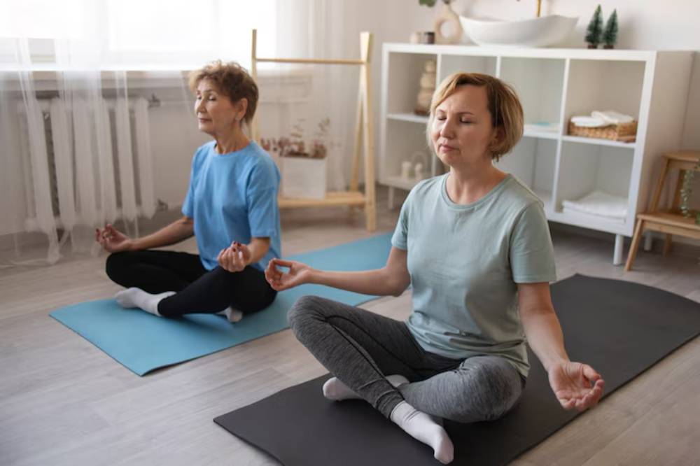 Two individuals meditate on yoga mats in a bright, serene room with natural light and wooden decor elements.
