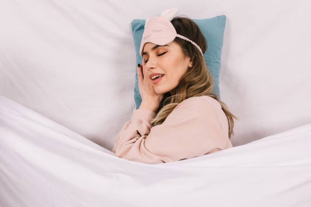 A cozy scene of a person lying in bed with a sleeping mask and soft pink pajamas, surrounded by white bedding and a teal pillow.