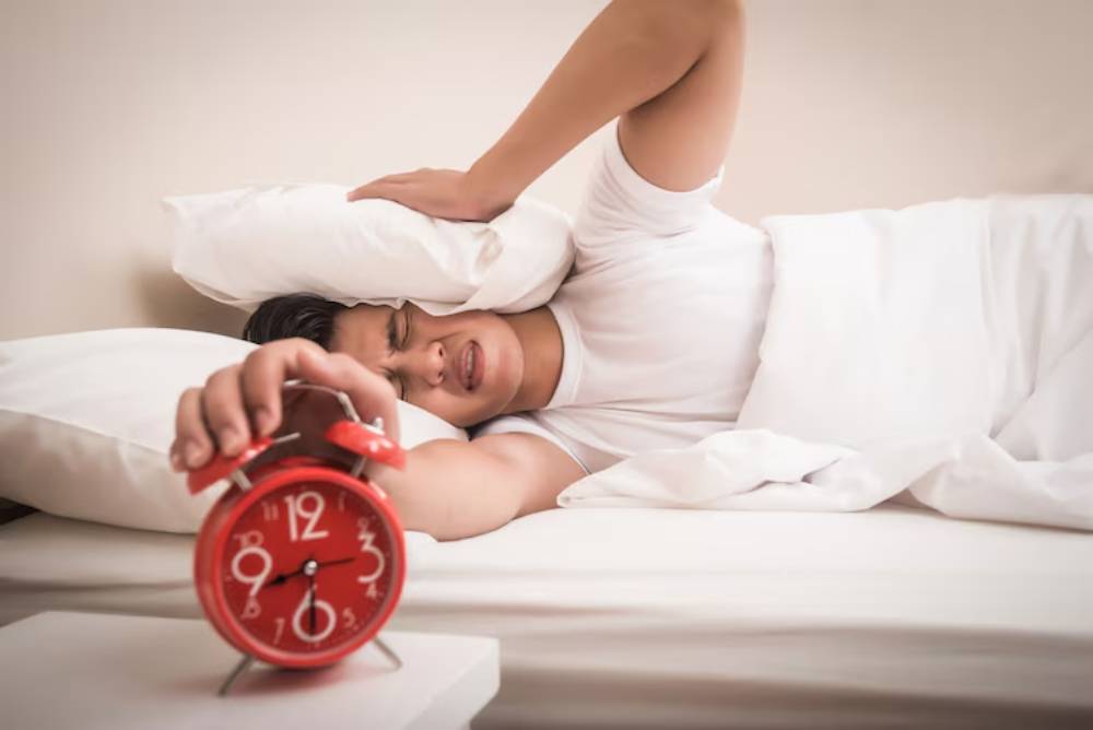 A person lies in bed, covered by white sheets, reaching to silence a red alarm clock on a bedside table.