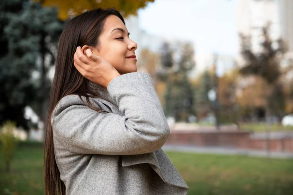 A woman in a gray coat stands outdoors, holding her hands to her ears, surrounded by trees and a park in autumn.