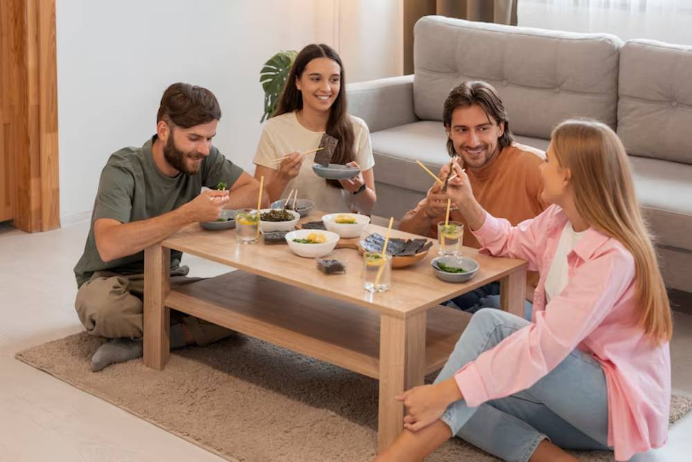 A group of four people enjoying a meal together at a wooden table, featuring various dishes and drinks in a cozy living room setting.