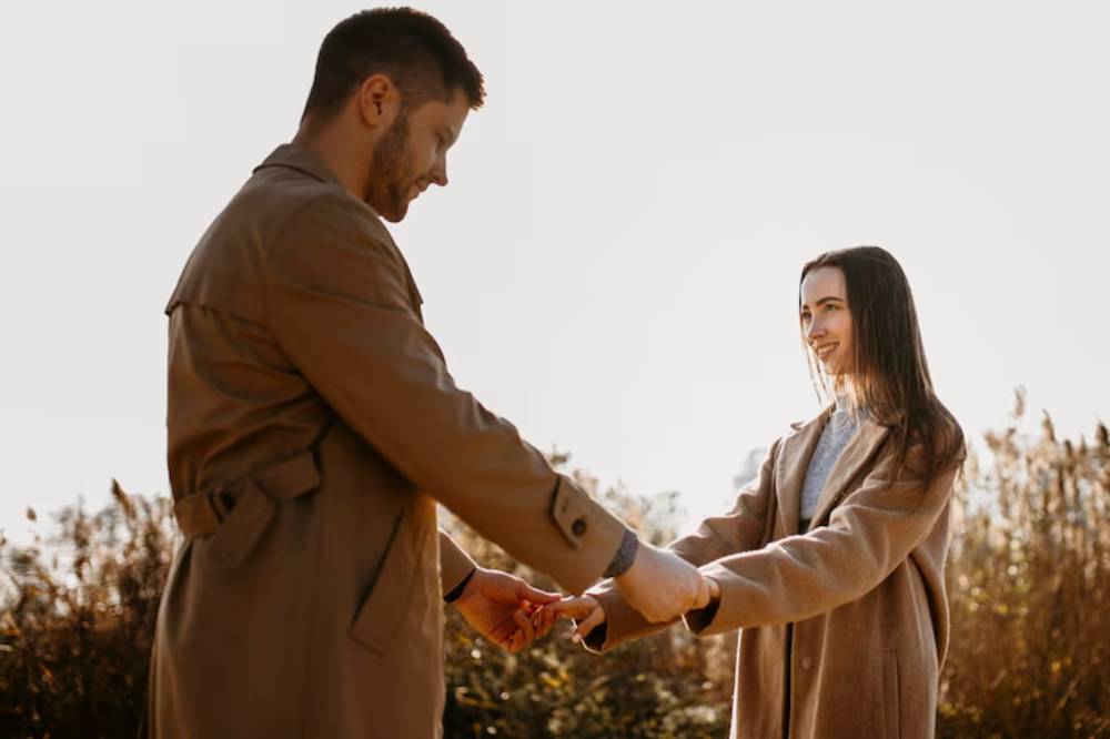 A couple stands outdoors, holding hands against a soft, blurred background of tall grass and a bright sky, conveying a sense of connection.