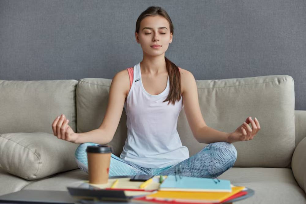 A person sits cross-legged on a couch, practicing mindfulness, with a coffee cup and colorful notebooks on the table in front.