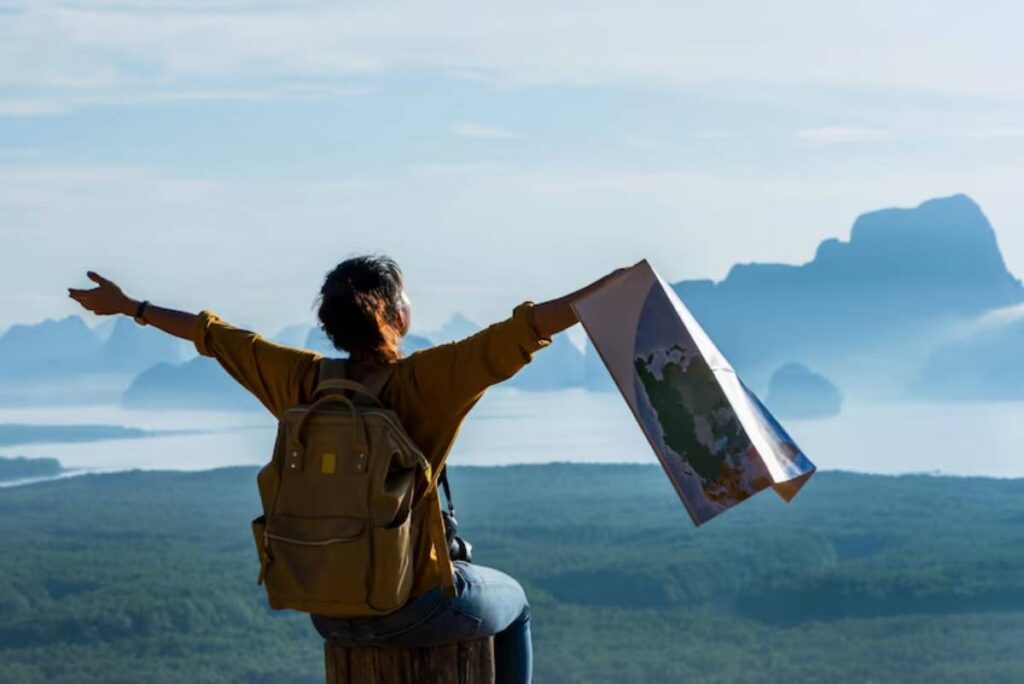 A woman with a backpack stands on a log, joyfully holding a map, overlooking a stunning mountainous landscape.