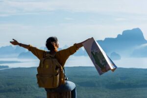 A woman with a backpack stands on a log, joyfully holding a map, overlooking a stunning mountainous landscape.