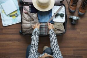 A person in a checkered shirt secures a packed suitcase with clothes, a hat, sunglasses, and a camera, surrounded by travel maps.