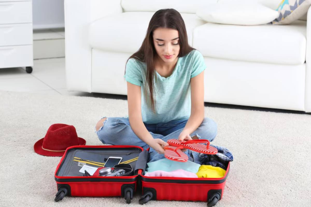 A person sitting on a carpet, sorting colorful clothes and accessories inside a red suitcase, with a red hat nearby.