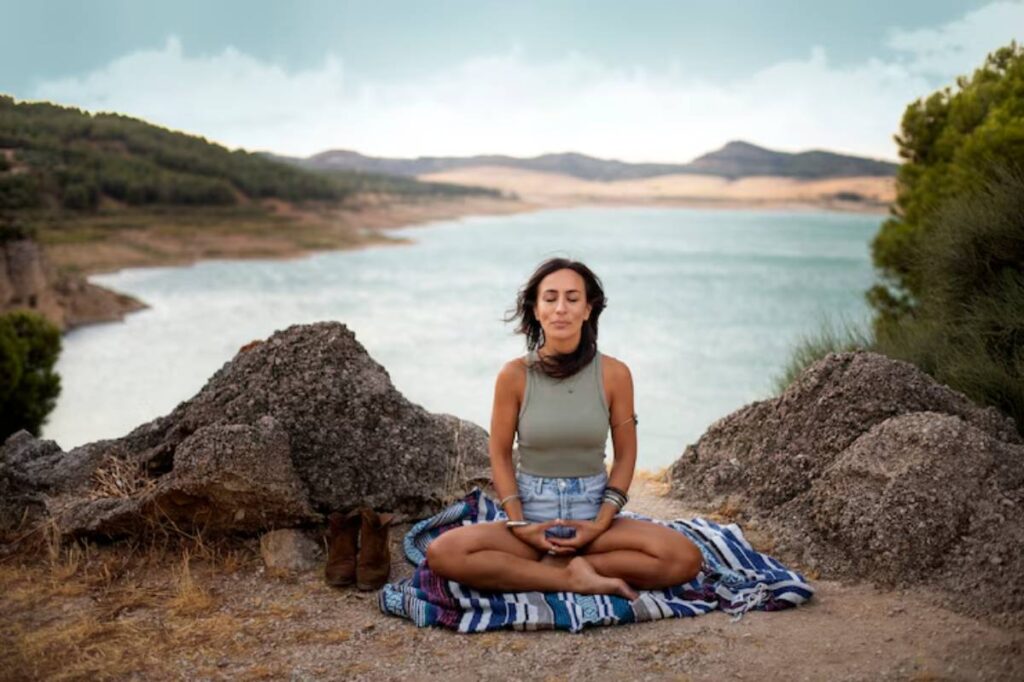 A person sits cross-legged on a colorful blanket by a serene lake, surrounded by rocky terrain and green hills under a cloudy sky.