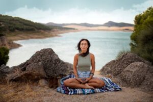 A person sits cross-legged on a colorful blanket by a serene lake, surrounded by rocky terrain and green hills under a cloudy sky.