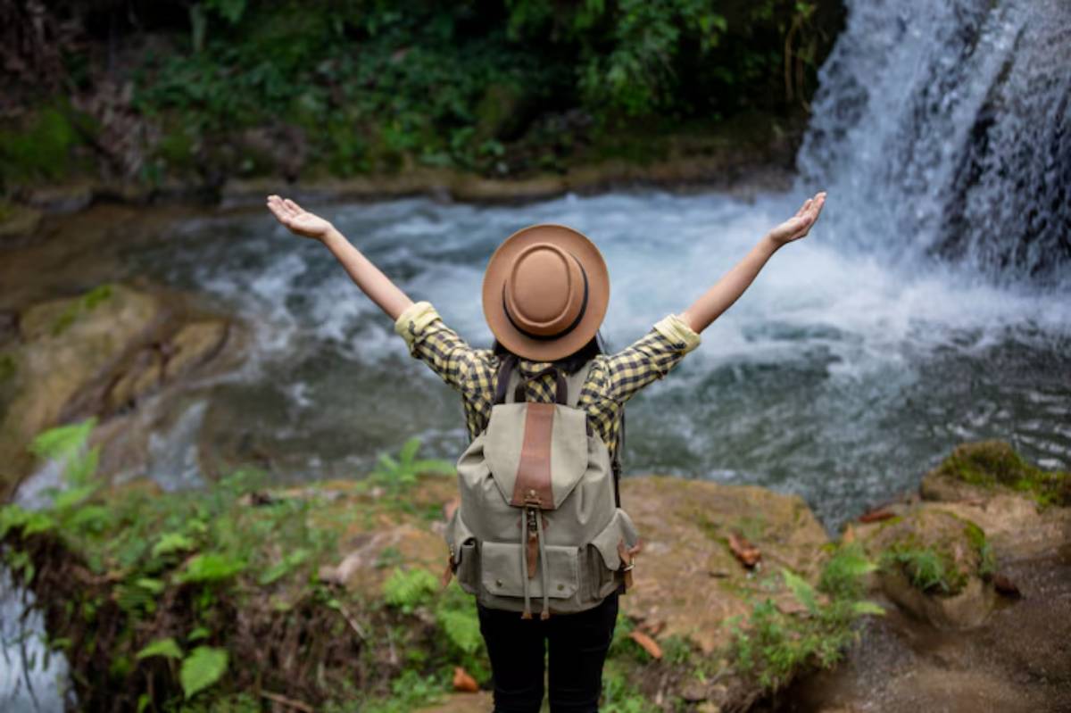 A woman in a plaid shirt and hat stands by a waterfall, arms raised, embracing nature with a joyful expression.