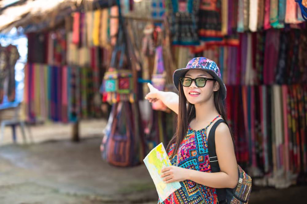 A person in a colorful patterned outfit holds a map and points, surrounded by vibrant textiles at a market.