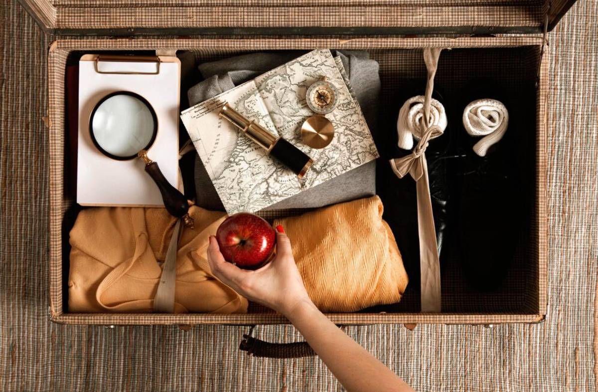A hand holds a red apple above a packed suitcase filled with clothing, a magnifying glass, a map, and other travel items.