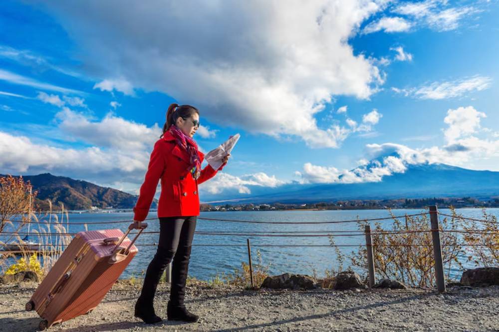 A woman in a red coat holds a map while standing by a lake with a suitcase, framed by mountains and a serene blue sky.