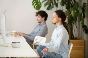 A woman meditates peacefully in a modern office setting, while a man works on a computer in the background, surrounded by greenery.