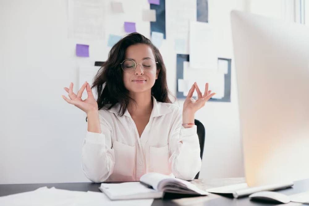 A person in a white shirt sits at a desk, practicing mindfulness with hands in a meditative gesture, surrounded by notes and a computer.