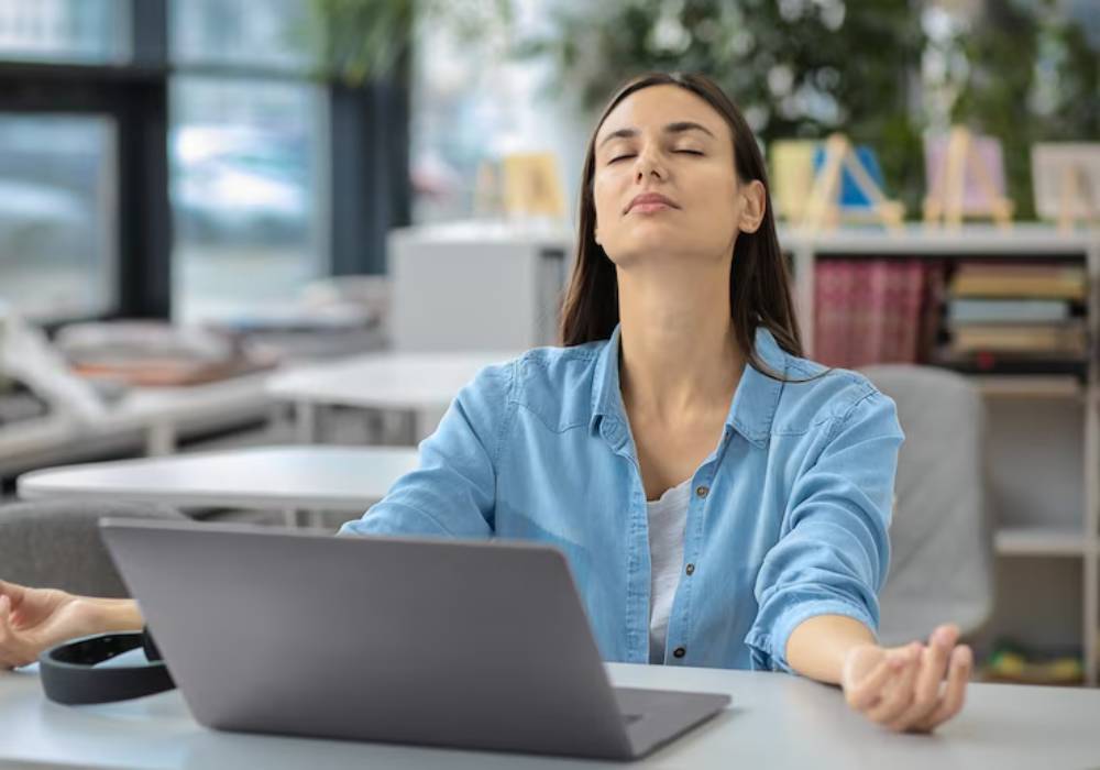 A woman in a blue shirt sits in a modern workspace, meditating with her hands resting on her knees, facing a laptop.
