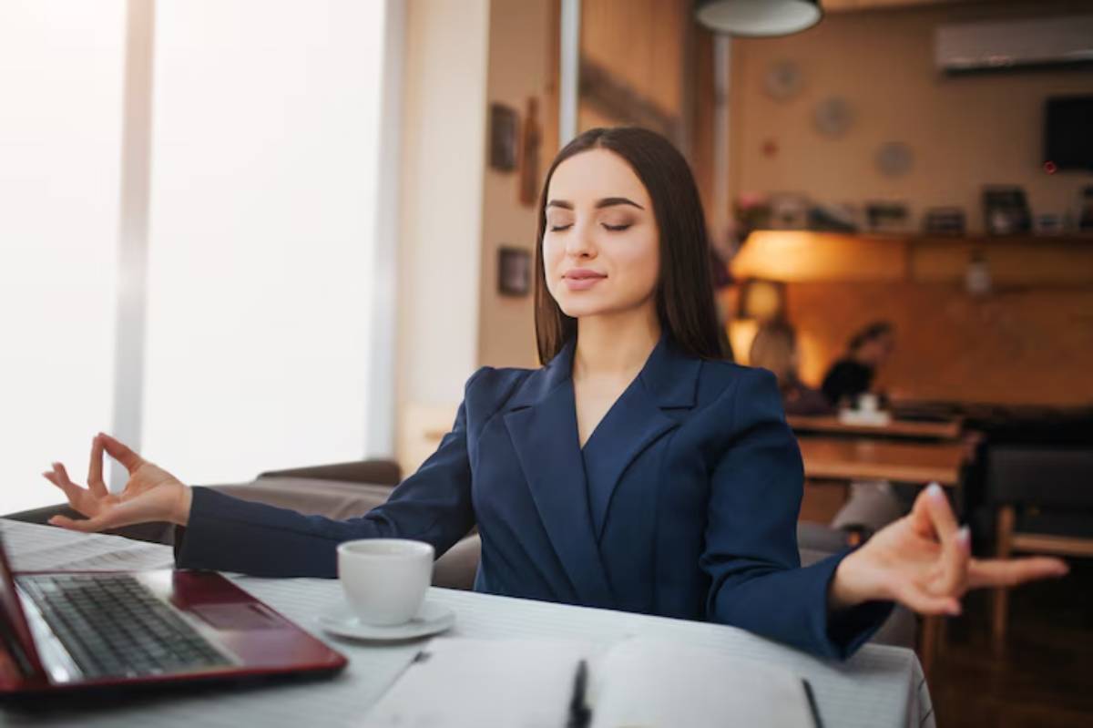 A woman in a dark blue blazer sits at a table, meditating with hands in a mudra, a laptop and coffee cup nearby.
