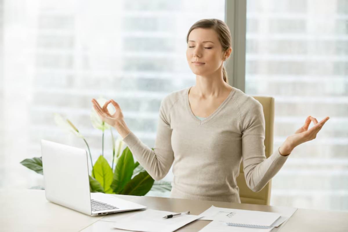 A woman practices mindfulness in an office setting, seated at a desk with a laptop and documents, surrounded by indoor plants.