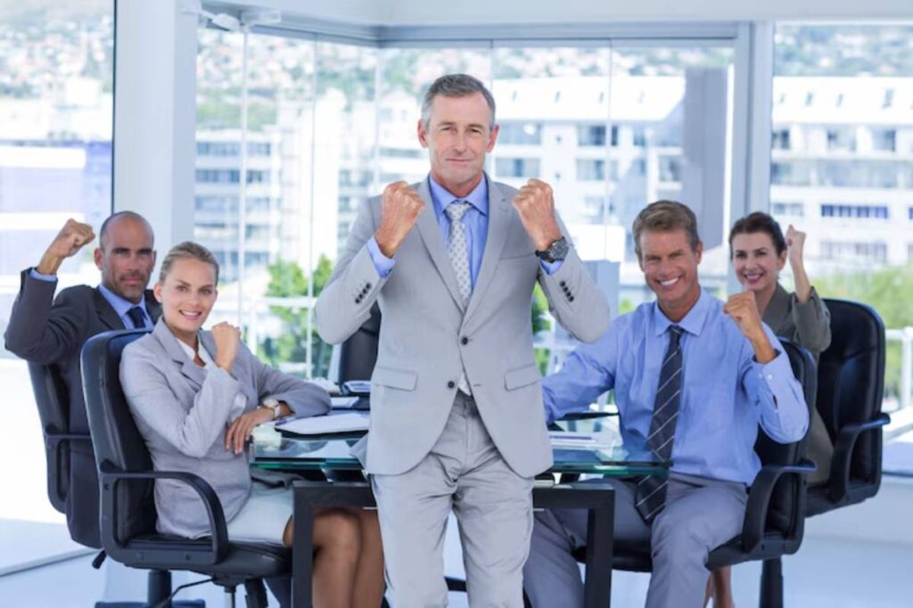A leader in a light gray suit celebrates passionately, while colleagues in a conference room cheer in excitement. Windows reveal a cityscape.